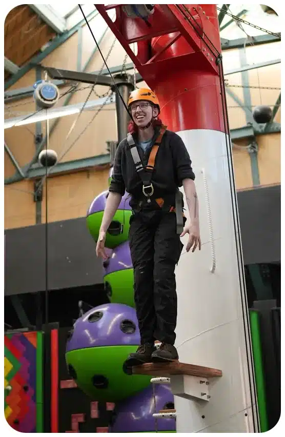 A young adult wearing a helmet and harness at the edge of a plank of wood about to take a leap of faith.
