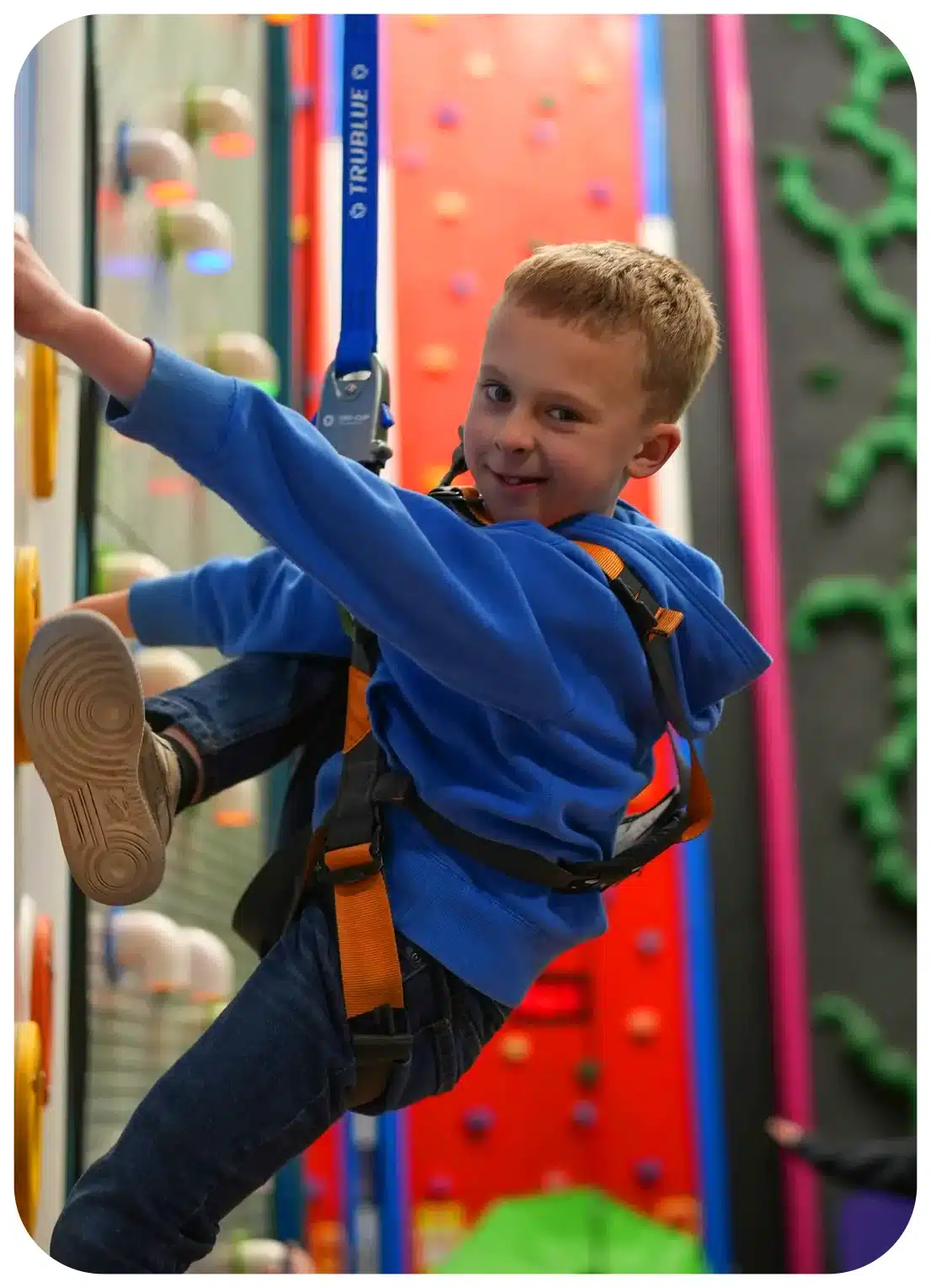 A child smiling at the camera whilst climbing up a climbing wall.